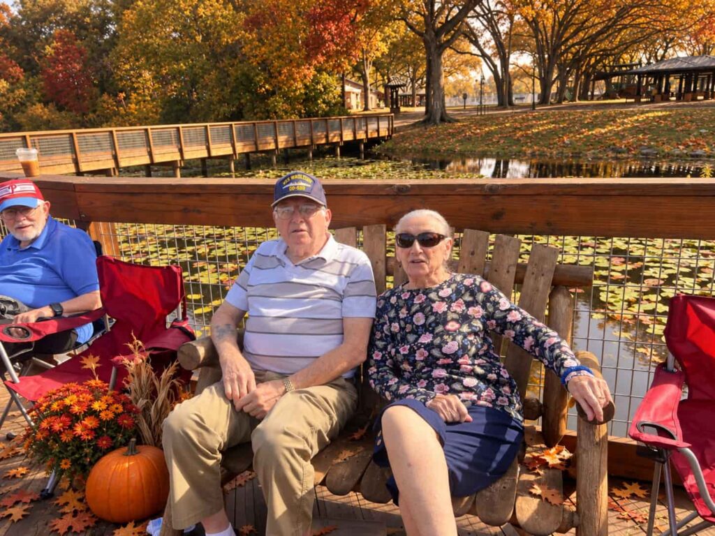 Three seniors sit on a wooden deck beside a pond with lily pads, surrounded by fall leaves, pumpkins, and orange flowers during an autumn outing at Madison at Oviedo.
