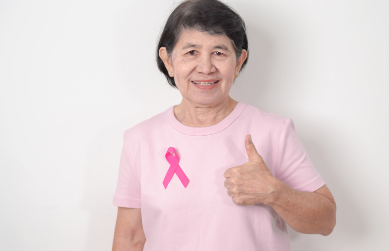 Smiling senior woman wearing a pink breast cancer awareness ribbon and giving a thumbs-up, promoting breast health awareness and preventive care for women over 70.